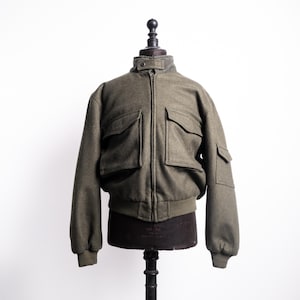 May include: An olive green bomber jacket with a ribbed hem and cuffs. The jacket has a zip-up front, a stand-up collar, and multiple front pockets. The jacket is displayed on a mannequin against a white backdrop.