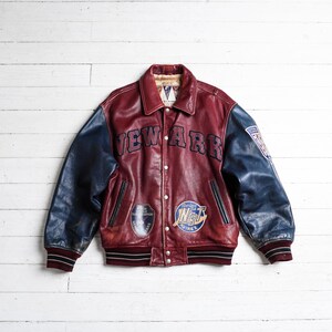 May include: A varsity-style leather jacket featuring a deep red body and dark blue sleeves. The front displays the text NEW ARK in dark blue lettering, along with two circular patches. The cuffs and hem are ribbed with black and white stripes.