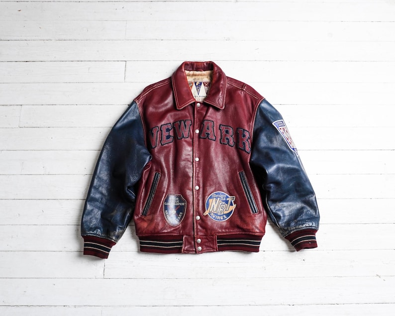 May include: A varsity-style leather jacket featuring a deep red body and dark blue sleeves. The front displays the text NEW ARK in dark blue lettering, along with two circular patches. The cuffs and hem are ribbed with black and white stripes.