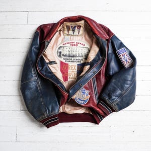 May include: A red and blue leather varsity jacket featuring a beige interior lining with Newark Bears 1949 Minor League Champions graphics. The jacket includes a State Final Champion patch on the sleeve and an Avirex USA logo on the inner lining.
