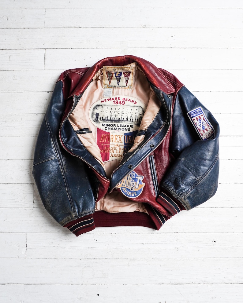 May include: A red and blue leather varsity jacket featuring a beige interior lining with Newark Bears 1949 Minor League Champions graphics. The jacket includes a State Final Champion patch on the sleeve and an Avirex USA logo on the inner lining.