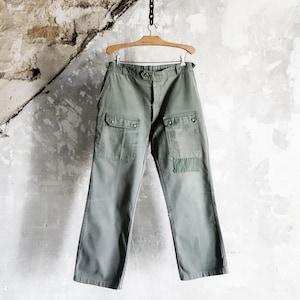 May include: Olive green cargo pants with two front pockets and two side pockets, hanging on a wooden hanger. The pants are made of a durable fabric and have a classic, utilitarian design. The background is a textured, off-white wall.
