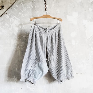 Op de afbeelding: Een gestreepte, korte broek, wit met verticale blauwe strepen, hangt aan een houten hanger. De broek heeft een geplooid zoom. De achtergrond is een getextureerde, gebroken witte muur.