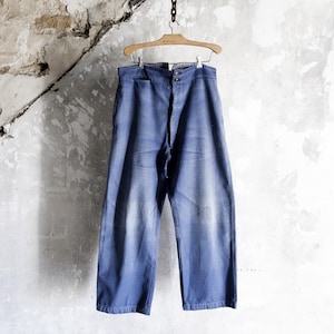 May include: Blue denim pants hanging on a wooden hanger against a textured, off-white wall. The pants have a relaxed fit, a button fly, and a drawstring. The denim has a faded appearance with some wear.