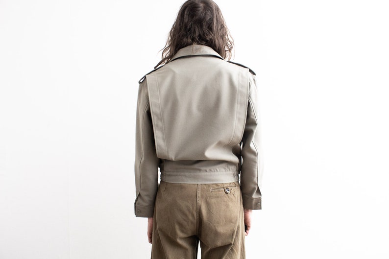 Pu&ograve; includere: Giacca beige chiaro dal design strutturato, con colletto, spalline e vita stretta. La giacca ha maniche lunghe ed &egrave; abbinata a pantaloni marroni. Il modello &egrave; visto da dietro.