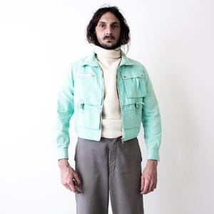May include: A light turquoise jacket with multiple pockets and a zipper closure. The jacket is worn over a cream-colored turtleneck sweater. The person is also wearing gray pants. The outfit is set against a white background.