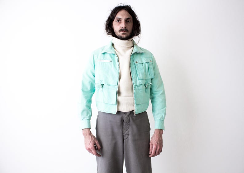 May include: A light turquoise jacket with multiple pockets and a zipper closure. The jacket is worn over a cream-colored turtleneck sweater. The person is also wearing gray pants. The outfit is set against a white background.
