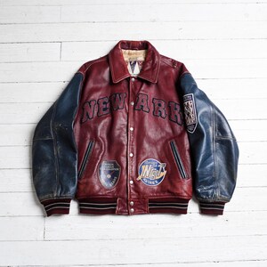 May include: A burgundy leather varsity jacket with navy blue sleeves, featuring the text NEW ARK across the chest. The jacket includes various embroidered patches, a button-front closure, and striped ribbed cuffs and hem in burgundy and black.