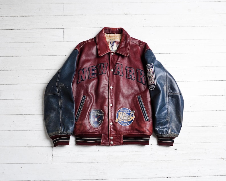 May include: A burgundy leather varsity jacket with navy blue sleeves, featuring the text NEW ARK across the chest. The jacket includes various embroidered patches, a button-front closure, and striped ribbed cuffs and hem in burgundy and black.