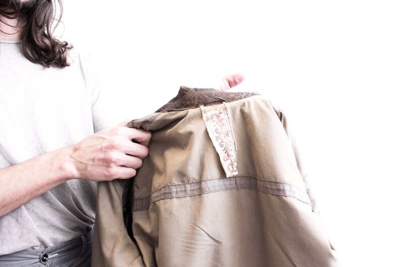 May include: A brown leather jacket with a beige lining and a decorative fabric tag. The jacket is being held by a person.