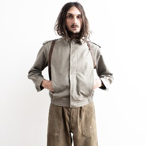 Pu&ograve; includere: Giacca verde oliva chiaro con chiusura a bottoni, cinghie in pelle marrone e orlo a coste. La giacca &egrave; abbinata a pantaloni verde oliva. Il modello &egrave; in piedi davanti a uno sfondo bianco.