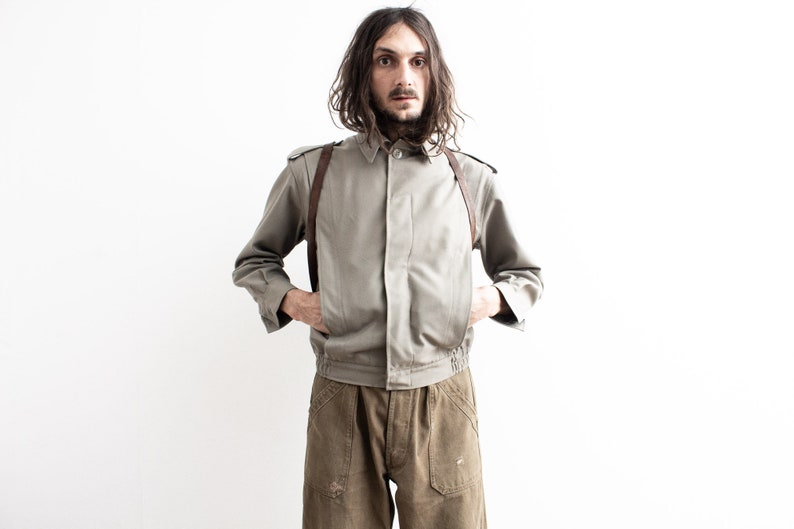 Pu&ograve; includere: Giacca verde oliva chiaro con chiusura a bottoni, cinghie in pelle marrone e orlo a coste. La giacca &egrave; abbinata a pantaloni verde oliva. Il modello &egrave; in piedi davanti a uno sfondo bianco.
