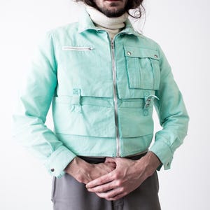 May include: A light turquoise jacket with multiple pockets and a silver zipper. The jacket has a collared neckline and long sleeves. The model is wearing a white turtleneck underneath. The jacket is a unique fashion piece.