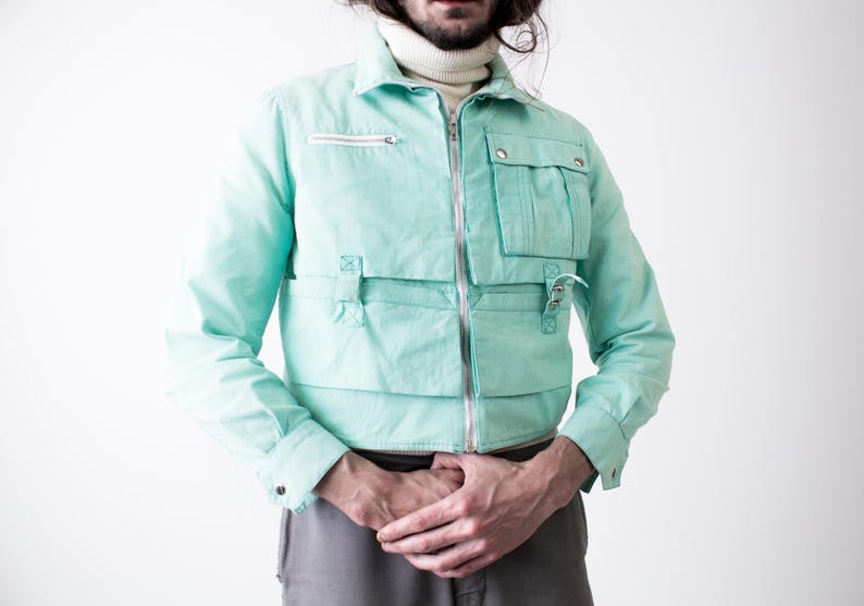 May include: A light turquoise jacket with multiple pockets and a silver zipper. The jacket has a collared neckline and long sleeves. The model is wearing a white turtleneck underneath. The jacket is a unique fashion piece.