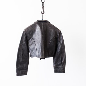 K&ouml;nnte beinhalten: Eine kurze, dunkelbraune Lederjacke mit umgelegtem Kragen, langen &Auml;rmeln und geschwungenen Nahtdetails auf der R&uuml;ckseite. Die Jacke h&auml;ngt an einem Metallhaken und zeigt eine verwitterte Textur sowie eine strukturierte Passform mit geradem Saum.