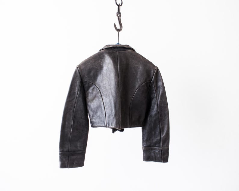 K&ouml;nnte beinhalten: Eine kurze, dunkelbraune Lederjacke mit umgelegtem Kragen, langen &Auml;rmeln und geschwungenen Nahtdetails auf der R&uuml;ckseite. Die Jacke h&auml;ngt an einem Metallhaken und zeigt eine verwitterte Textur sowie eine strukturierte Passform mit geradem Saum.