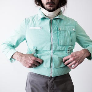 May include: A light turquoise jacket with multiple pockets and a zipper closure. The jacket has a collared neckline and long sleeves. The model is wearing a white turtleneck underneath. The jacket has silver-colored hardware.