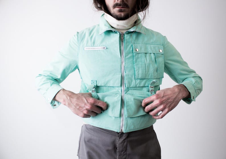 May include: A light turquoise jacket with multiple pockets and a zipper closure. The jacket has a collared neckline and long sleeves. The model is wearing a white turtleneck underneath. The jacket has silver-colored hardware.