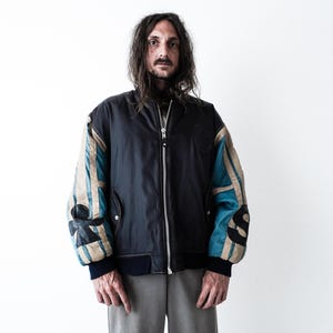 May include: A black bomber jacket featuring a central silver zipper, long sleeves with beige and teal vertical panels, and black circular patches on the elbows. The jacket has a ribbed black collar, cuffs, and hem, with a solid black front panel.