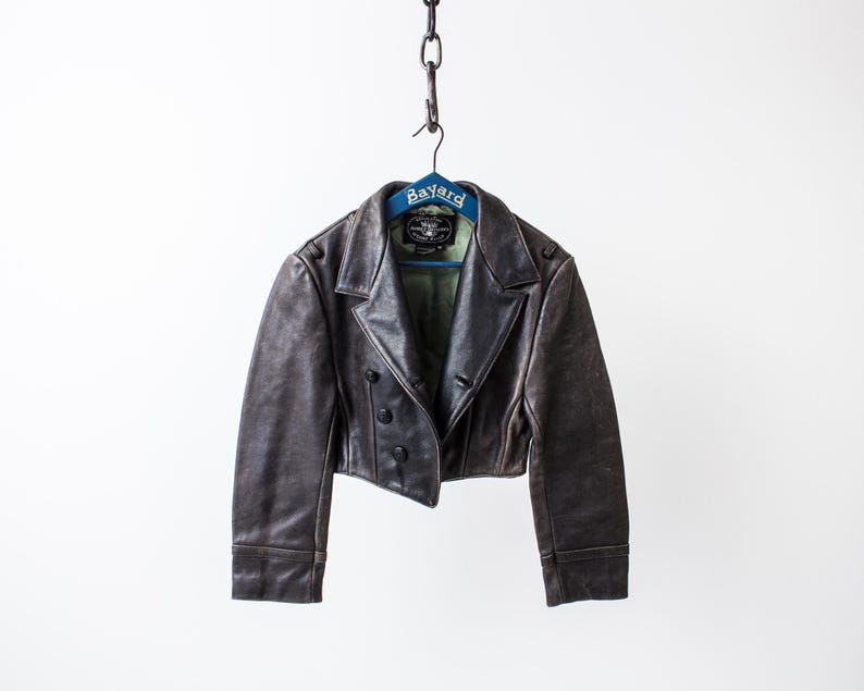 K&ouml;nnte beinhalten: Eine kurze, dunkelbraune Lederjacke mit breitem Reverskragen, langen &Auml;rmeln und einer zweireihigen Knopfleiste mit vier schwarzen Kn&ouml;pfen. Die Jacke h&auml;ngt an einem blauen Kleiderb&uuml;gel mit der Aufschrift Bayard und zeigt ein hellgr&uuml;nes Innenfutter.
