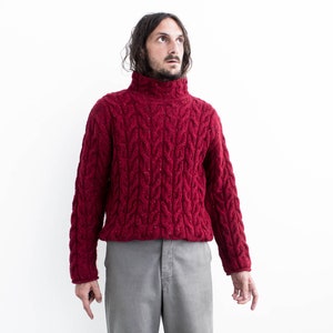 Op de afbeelding: Een man draagt een rode kabelgebreide trui met een hoge hals en een grijze broek. Hij staat voor een witte achtergrond.