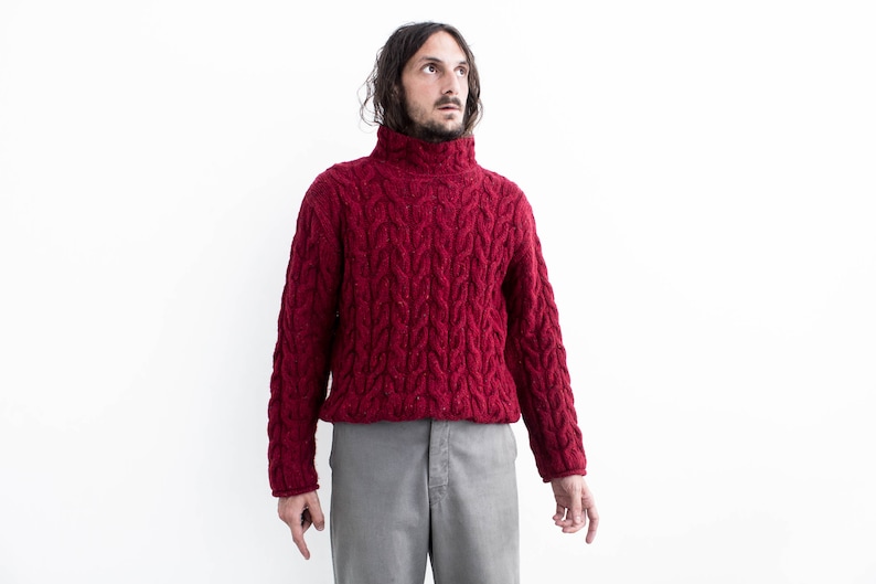 Op de afbeelding: Een man draagt een rode kabelgebreide trui met een hoge hals en een grijze broek. Hij staat voor een witte achtergrond.