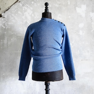 Könnte beinhalten: Ein blauer Pullover mit hohem Ausschnitt und langen Ärmeln, präsentiert auf einer Vintage-Schneiderpuppe. Der Pullover hat dunkle Knöpfe auf der Schulter. Das Kleidungsstück ist aus einem weichen, strukturierten Material gefertigt.