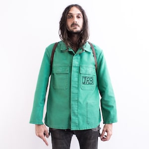 Peut inclure: Un homme portant une veste de travail verte avec une sangle en cuir marron et un patch noir qui dit "JRB" sur le devant. Il porte un jean bleu et a les cheveux longs et bruns.
