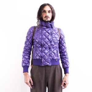 Puede incluir: Una chaqueta acolchada de color morado con cuello alto y cierre de cremallera. La chaqueta tiene un patrón de diamante y la lleva puesta una persona con el pelo largo y oscuro.