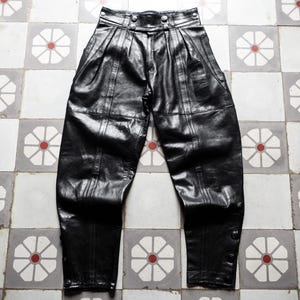 Puede incluir: Pantalón de cuero negro de cintura alta con pliegues. El pantalón tiene un corte ajustado y una pierna recta.