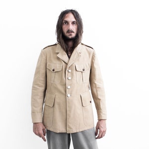 May include: A man wearing a beige safari jacket with four buttons and two flap pockets. The jacket has a collar and long sleeves.