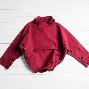 Könnte beinhalten: Ein weinrotes Oberteil mit hohem Kragen und langen Ärmeln. Es hat schwarze Knöpfe am Kragen, an den Manschetten und an den Seiten. Das Kleidungsstück ist aus einem weichen Stoff gefertigt und wird auf einer weißen Holzoberfläche präsentiert.