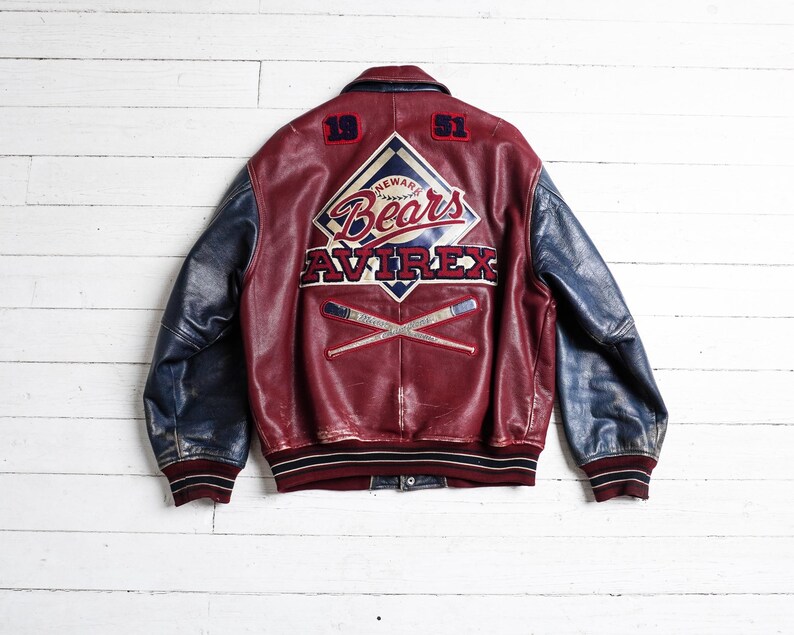 May include: A maroon and blue leather varsity jacket featuring the text Newark Bears and Avirex on the back. The back design includes the numbers 19 and 51, a diamond-shaped logo, and two crossed baseball bats. The jacket has a ribbed collar and cuffs.