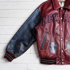 May include: A maroon leather varsity jacket with navy blue leather sleeves. The jacket features the text NEW A in dark blue lettering, a Newark Bears Minor League Club Champion patch, a second circular patch, and striped maroon and navy cuffs.
