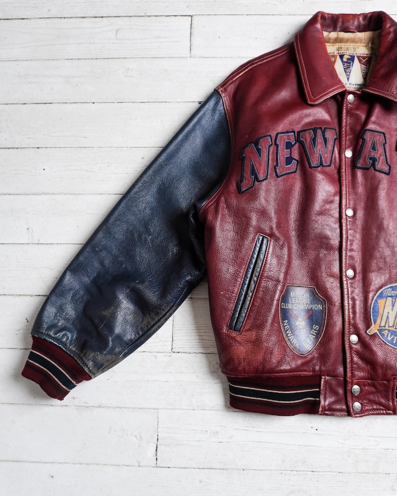May include: A maroon leather varsity jacket with navy blue leather sleeves. The jacket features the text NEW A in dark blue lettering, a Newark Bears Minor League Club Champion patch, a second circular patch, and striped maroon and navy cuffs.