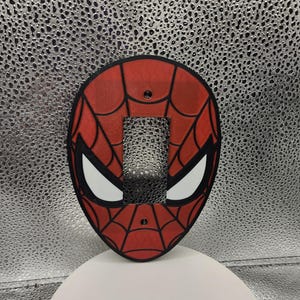 May include: A red and black light switch cover plate featuring a spiderweb pattern and large white eye shapes. The oval-shaped cover has a rectangular cutout in the center for a toggle switch and two small screw holes at the top and bottom.