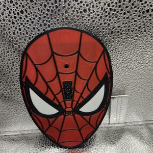 Könnte beinhalten: Eine rote und schwarze Spider-Man-Maske mit einem markanten Spinnennetz-Muster auf der Oberfläche. Die Maske hat große, weiße, mandelförmige Augenlinsen mit schwarzer Umrandung und ein kleines, schwarzes, rechteckiges Detail über dem Nasenbereich.