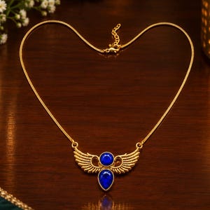 May include: A gold-toned necklace featuring a central pendant with two stylized wings extending outward. The pendant is accented with a circular blue stone at the top and a larger, teardrop-shaped blue stone positioned directly below it.