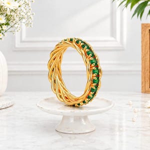 Pode incluir: Um bracelete de ouro com um design de metal torcido e uma fileira de pedras preciosas redondas de um verde vibrante engastadas ao longo da borda interna. O bracelete tem um acabamento metálico polido e uma estrutura circular detalhada.
