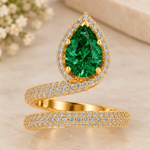 May include: A gold bypass ring featuring a pear-shaped green center stone surrounded by a halo of small white stones. The band is a wrap-around style, fully encrusted with multiple rows of tiny white stones that extend from the central setting.