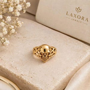 May include: A gold ring featuring a detailed skull design with an intricate, web-like band. The ring is displayed on a textured stone surface next to a white box with the text LAXORA JEWELS and a small floral logo printed on the top.