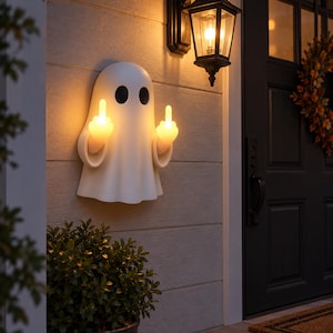 Peut inclure: Une applique murale en forme de fantôme blanc avec deux yeux ovales noirs. Le fantôme a deux bras levés, les majeurs tendus et illuminés d'une lueur jaune chaude, créant un effet lumineux brillant depuis les mains contre le mur.