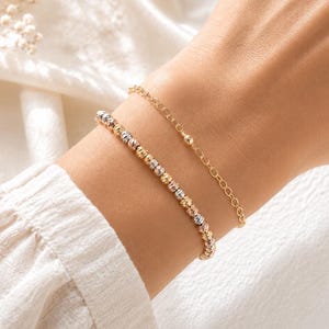Puede incluir: Dos pulseras doradas se muestran en una muñeca. Una es una cadena delicada con un pequeño dije de bola. La otra es una pulsera de cuentas con cuentas tricolores en oro, plata y oro rosa. Las pulseras están sobre un fondo blanco suave.