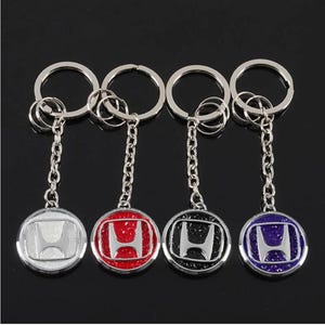 May include: Four circular metal keychains featuring the silver Honda H logo centered on a textured background. Each keychain includes a silver split ring and a short chain. The background colors for the logos are white, red, black, and purple.