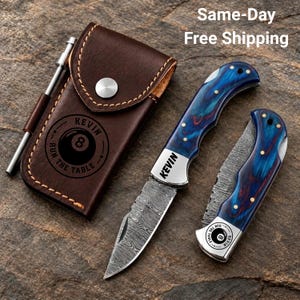 May include: A brown leather pouch with a silver snap button and a printed 8-ball graphic with the text KEVIN RUN THE TABLE, alongside a metal pen and two folding knives with blue wood-grain handles and patterned steel blades.