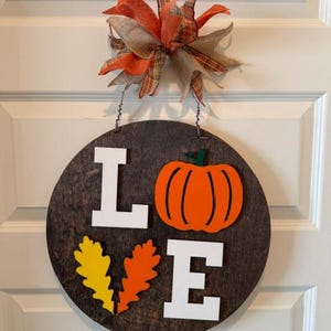 Puede incluir: Un letrero circular de madera marrón oscuro con la palabra LOVE en letras blancas, con una calabaza naranja reemplazando la letra O y dos hojas de roble amarillas y naranjas reemplazando la letra V. Un lazo de tela multicolor decora la parte superior.