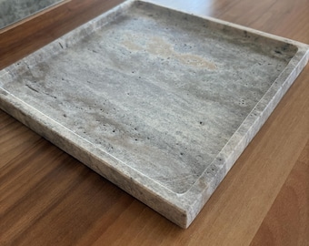 Könnte beinhalten: Ein quadratisches Steintablett mit einem natürlichen grau-beigen Marmormuster und einer porösen, strukturierten Oberfläche. Das Tablett hat einen erhöhten Rand und einen flachen Boden, der die organischen Variationen und Aderungen zeigt.
