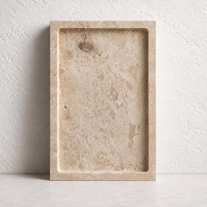 Puede incluir: Una bandeja rectangular hecha de piedra travertino beige, con una superficie lisa y pulida que presenta texturas porosas naturales y vetas minerales variadas en tonos bronceado y crema por todo el material, con un diseño plano y abierto.