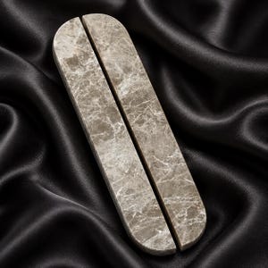 May include: A pair of elongated, pill-shaped stone objects featuring a polished, light brown and gray marble surface with intricate white veining patterns. The two pieces are positioned side-by-side, displaying smooth, rounded ends and flat edges.