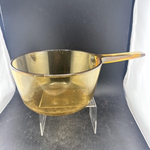 May include: A round, amber-coloured glass saucepan featuring a single, integrated glass handle extending from the side. The vessel has a smooth, translucent finish and a wide, open top, showcasing a simple, functional design for kitchen use.